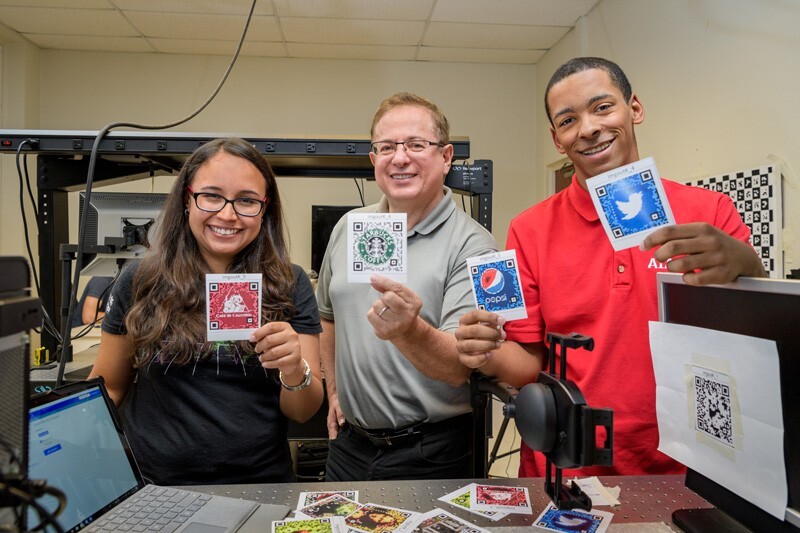 From left to right, UD doctoral student Karelia Pena; Gonzalo Arce, Charles Black Evans Professor of Electrical and Computer Engineering; and David Miller, a student at Mount Pleasant High School in Wilmington, Delaware, show the QR codes they have studied using sophisticated optical equipment in a laboratory in UD's Evans Hall. 