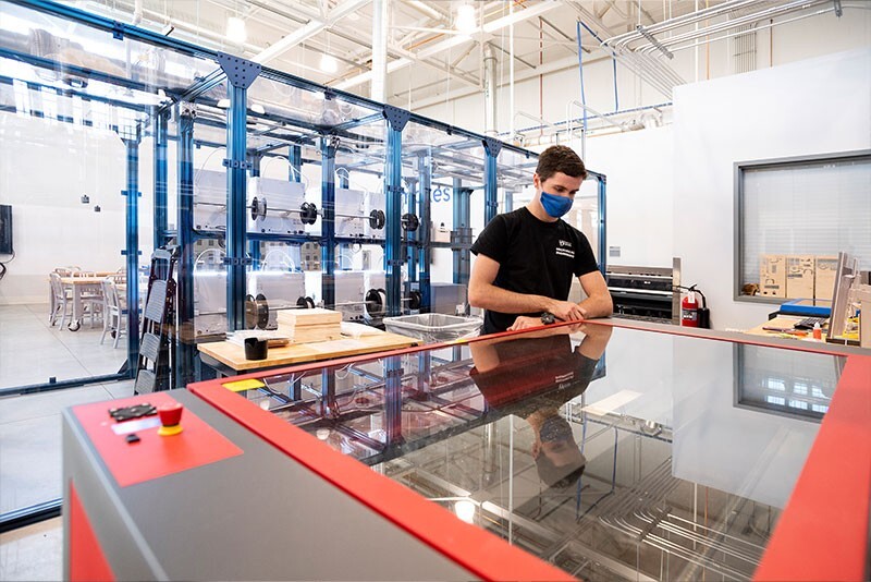A student uses a laser cutting machine while standing in front of a bank of 3D printers.