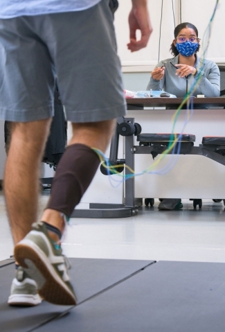 Foreground, a person walking on a treadmill with sensors on their leg. Hannah Armstrong is in the background providing direction to the walker.