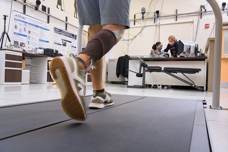 Person walking on a treadmill with wides and sensors on their leg. In the background, two researchers lean over a laptop.