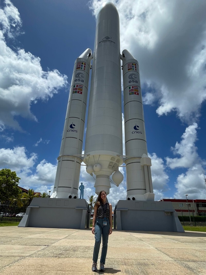 Elaine Stewart is standing in front of a mockup of the Ariane 5 rocket that will carry the telescope into orbit. The rocket has a central payload with two rocket boosters on each side.