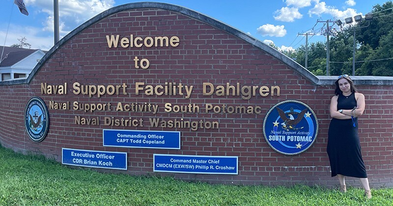 Hannah Epstein standing in front of a brick sign for the Naval Support Facility Dahlgren.