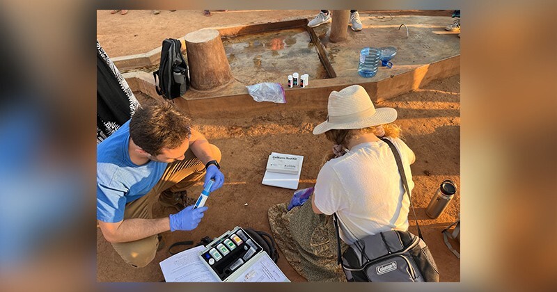 Jerel Okonski (left) and Hannah Bockius sit on the ground while conducting water tests with a hand operated pump in the background.
