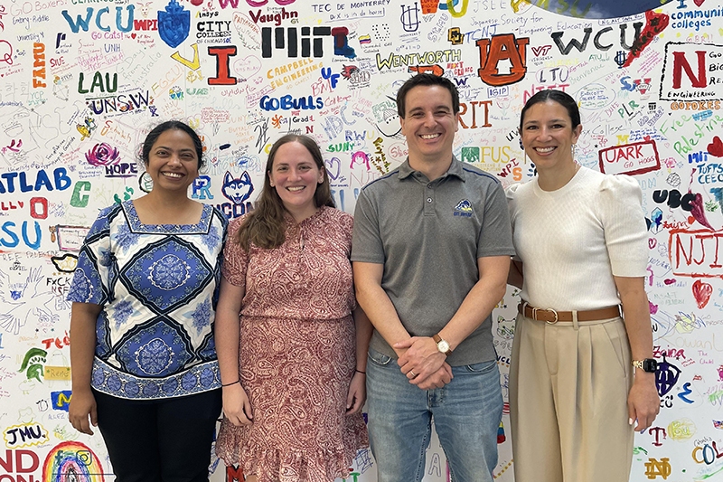 Four people stand side by side and smile at the camera in front of a wall covered with colorful drawings, words and university logos.