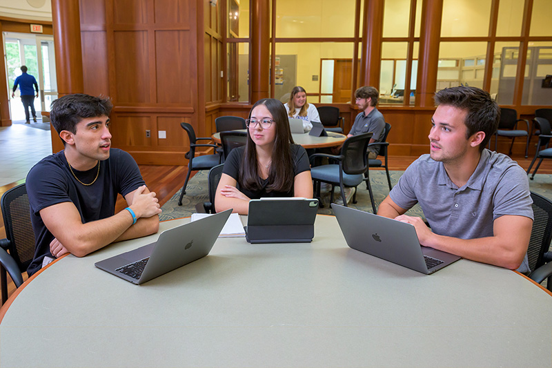Three students sit at a roundtable talking with laptops open in front of them.