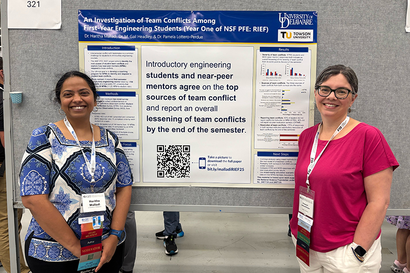 Two women stand and smile in front of a research poster. The woman on the left wears a blue patterned top and black pants, and the woman on the right wears glasses, a pink shirt and white pants. The poster behind them includes text, charts and a QR code.