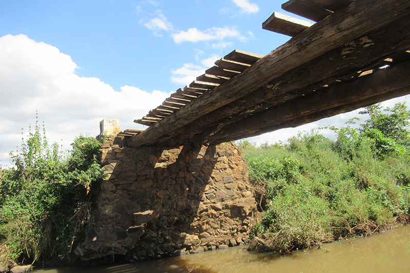 A deteriorating wooden plank bridge over a river. some of the cross planks are missing.